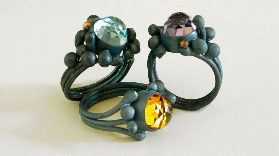 Drei grau-schwarze Ringe mit jeweils einem Stein in der Mitte, in den Farben: Blau, lila und gelb